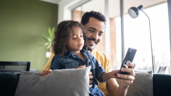 父亲和孩子坐在一起微笑而看智能手机。”loading=